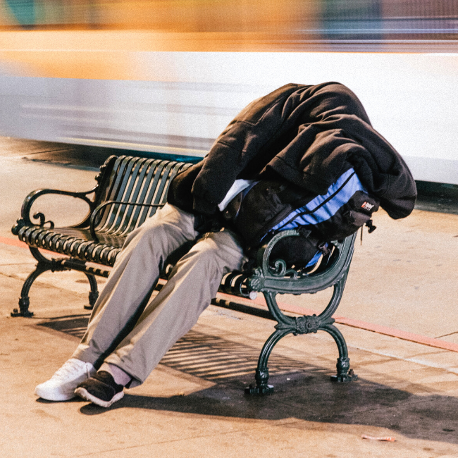 A homeless man sat on a bench with a duvet covering his upper body and head